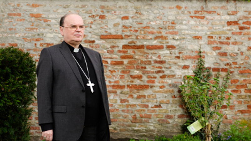 Bischof Bertram Meier steht in einem Garten vor einer Ziegelwand