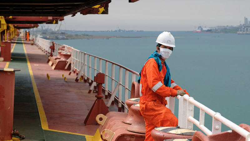 Seemann steht mit Mundschutz auf einem Schiff im Hafen