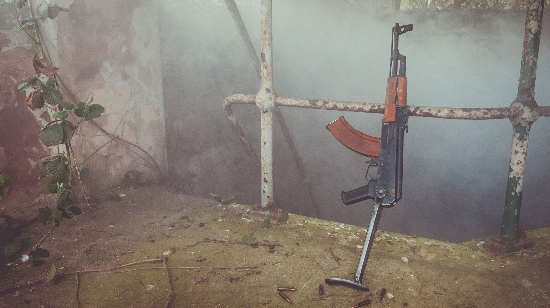 Ein Gewehr steht an einem rostigen Geländer angelehnt inmitten von Nebelschwaden.