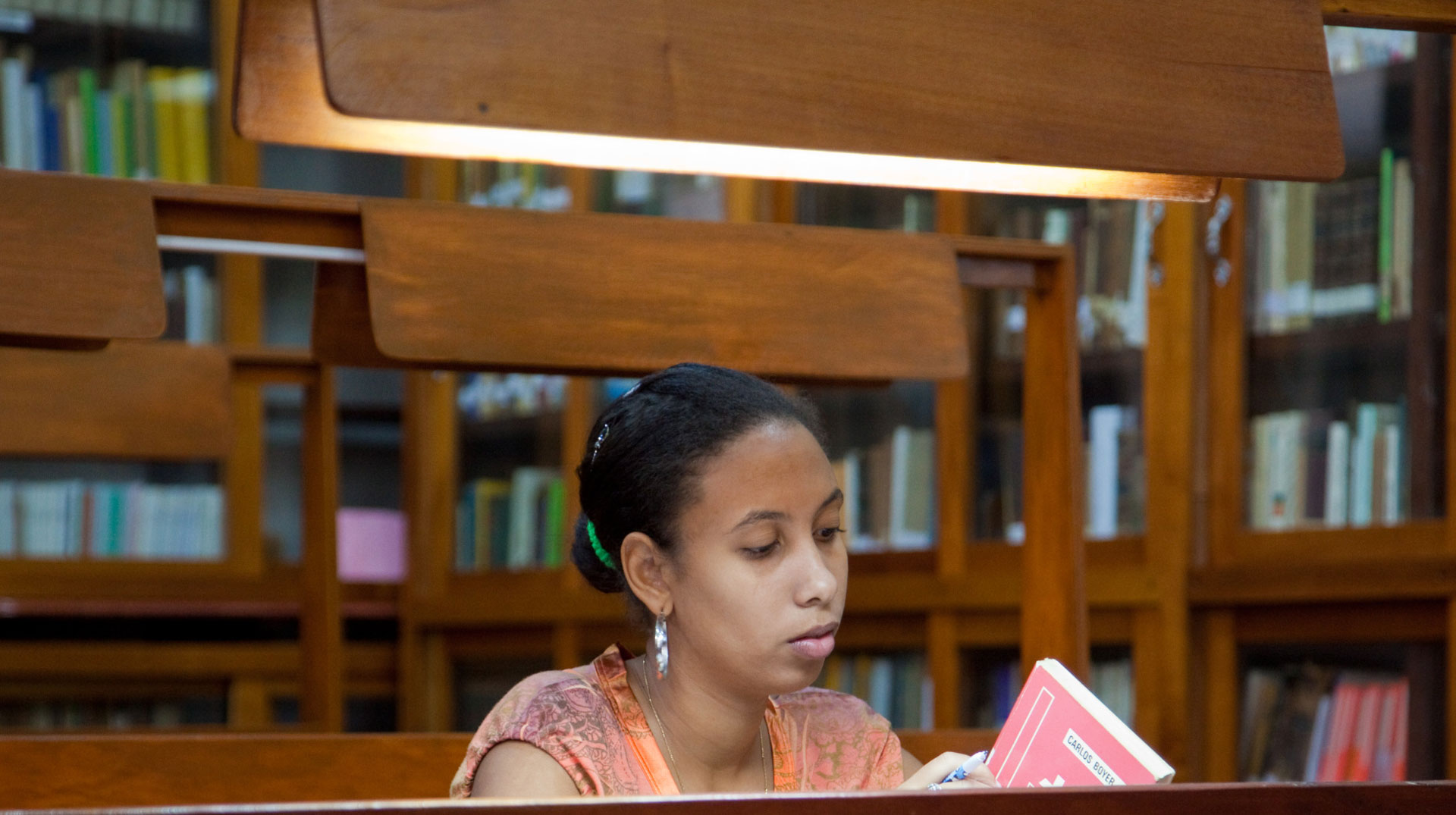 Eine Frau sitzt unter einer Lampe an einem schweren Bibliothekstisch. Im Hintergrund Regale mit Büchern.