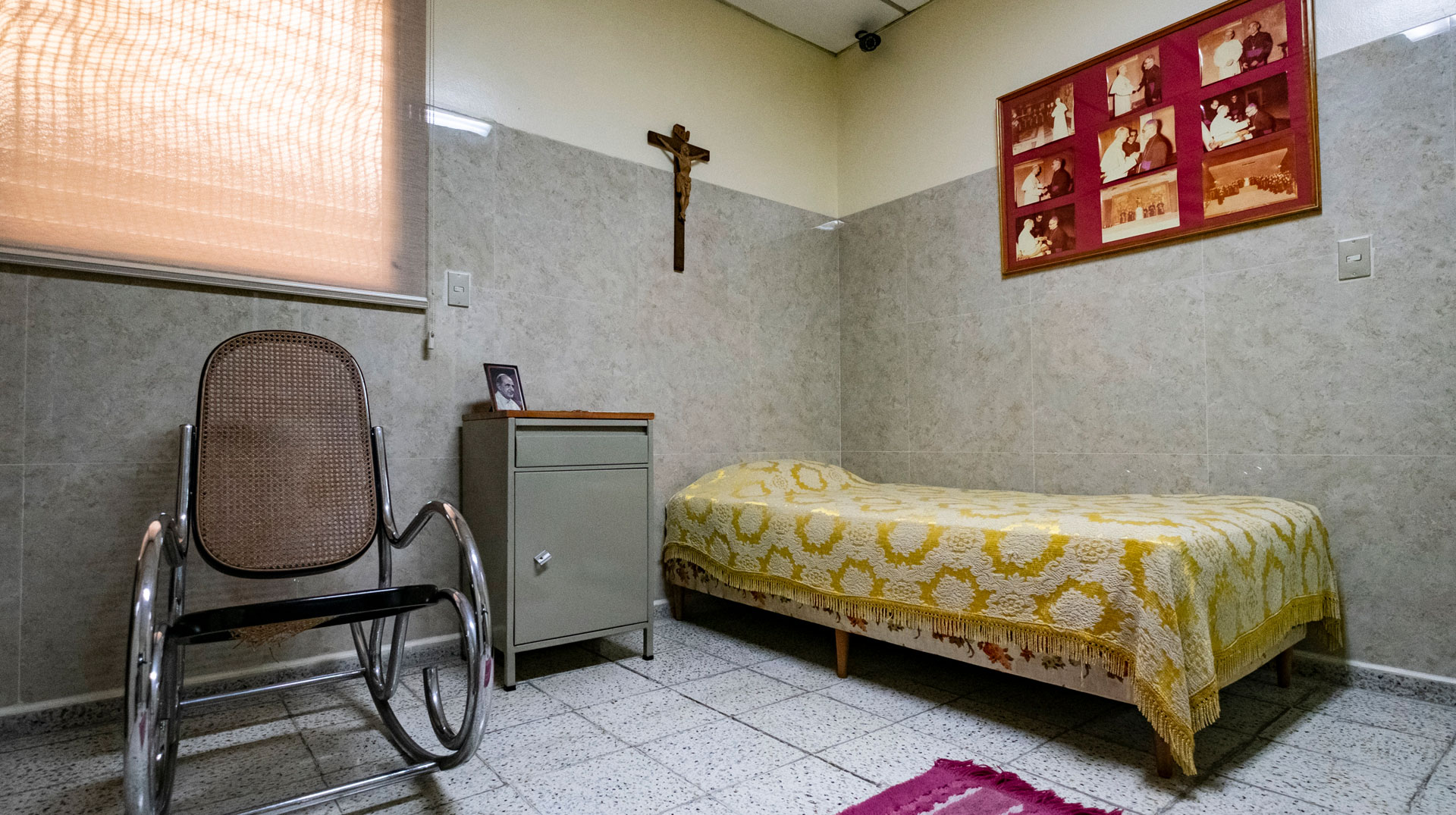 In einem kleinen, gefliesten Raum steht ein einfacher Schaukelstuhl, ein kleiner Metallschrank und ein einfaches Bett. An der Wand hängt ein Kreuz und ein Bilderrahmen mit Fotos des heiligen Óscar Romero bei Papstaudienzen.