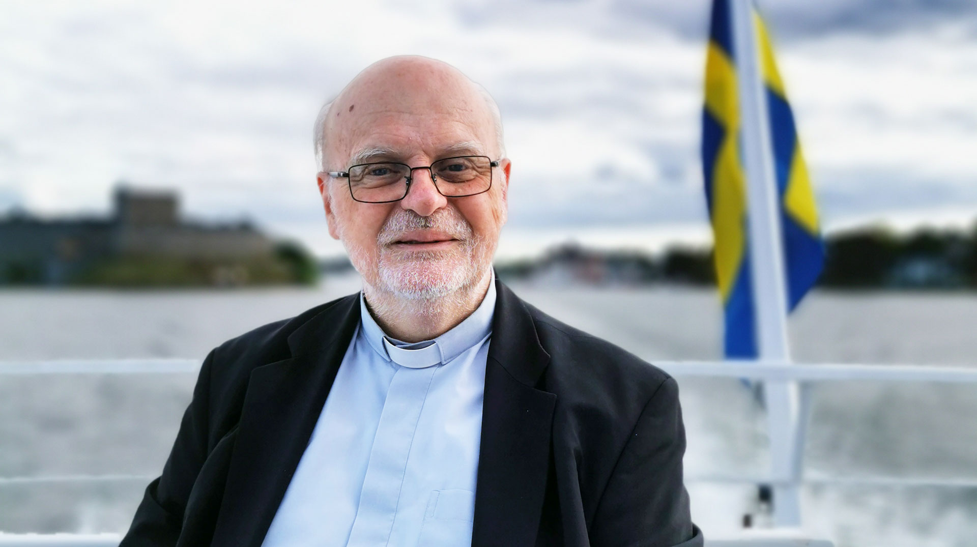 Kardinal Anders Arborelius sitzt auf einem Boot, im Hintergrund die schwedische Flagge