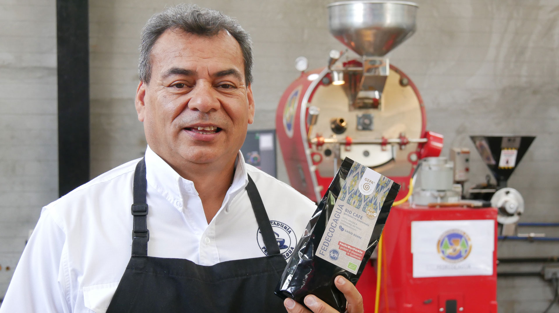 Luis Alvarado, Röstmeister beim GEPA-Kaffeepartner FEDECOCAGUA in Guatemala, zeigt eine Kaffeeverpackung und steht vor der Röstmaschine.
