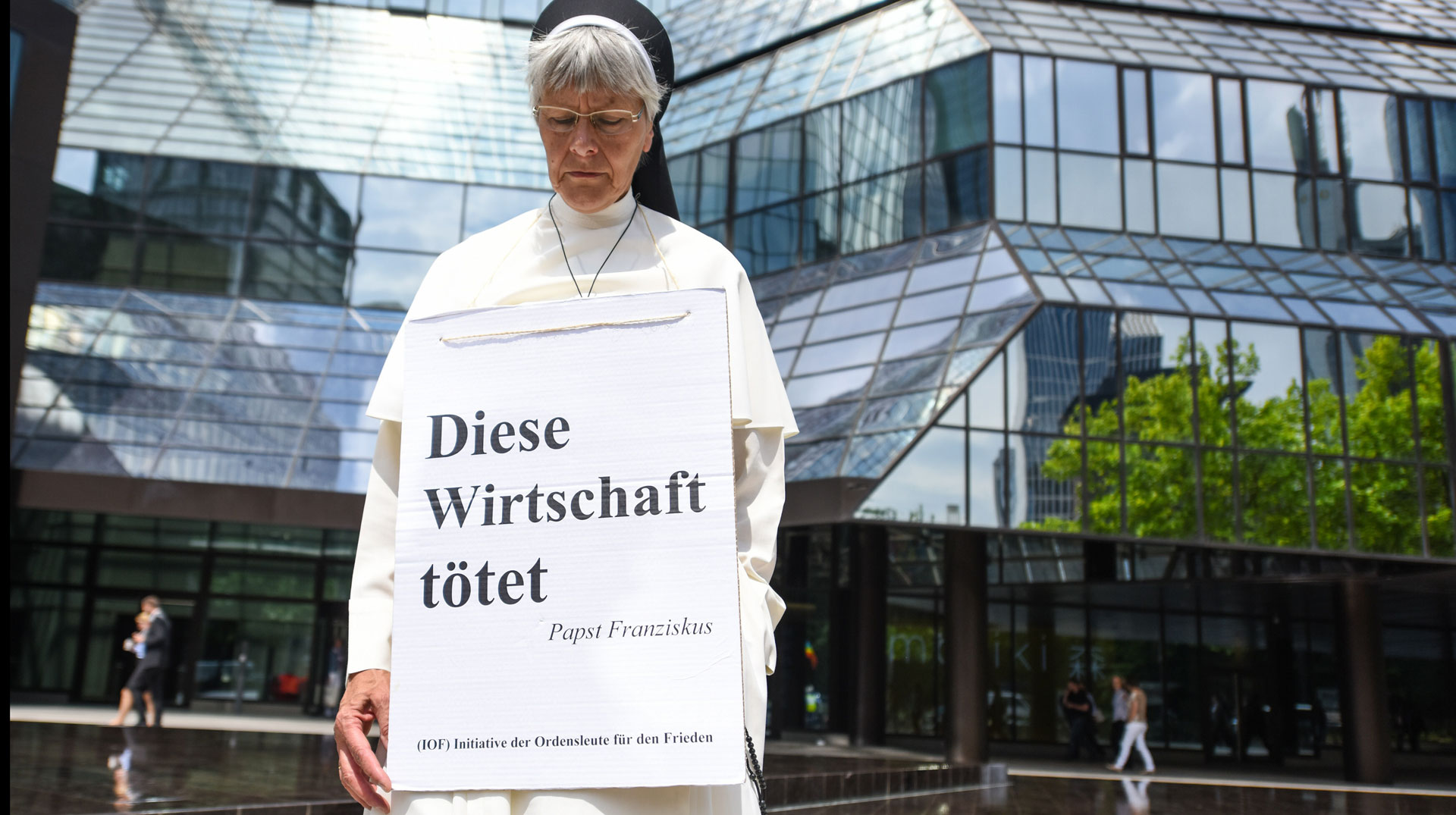 Ordensfrau steht vor dem Hauptsitz der Deutschen Bank AG in Frankfurt. Um ihr Hals ein Plakat mit der Aufschrift "Diese Wirtschaft tötet".