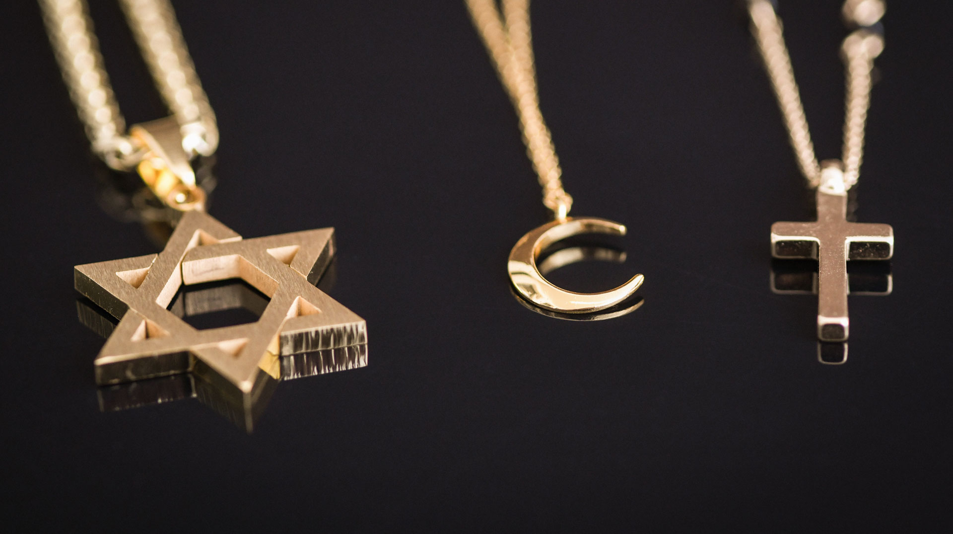 Ein goldfarbener Davidstern, ein Halbmond und ein Kreuz als Anhänger an drei Ketten liegen nebeneinander auf schwarzem Untergrund am 16. Dezember 2019 in Bonn. Symbolbild interreligiöser Dialog Religionen Weltreligionen