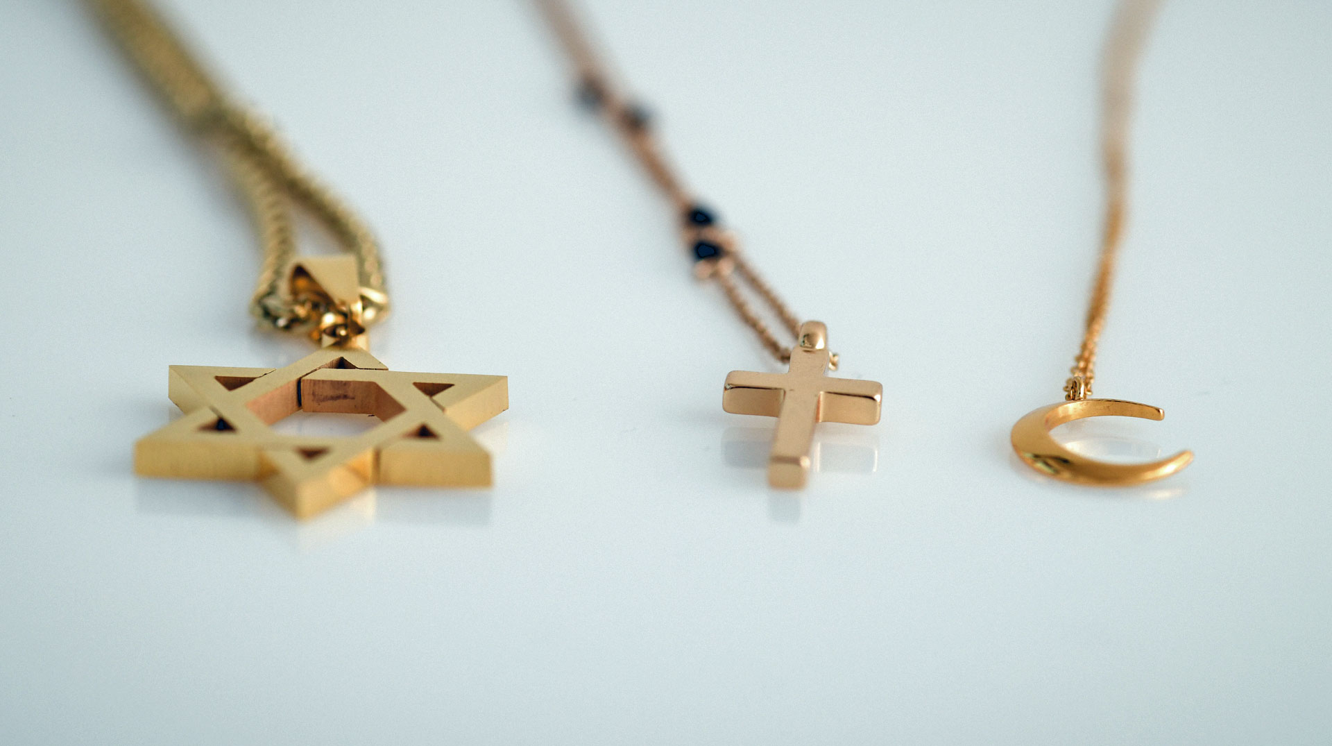 Ein goldfarbener Davidstern, ein Kreuz und ein Halbmond als Anhänger an drei Ketten liegen nebeneinander auf weißem Untergrund