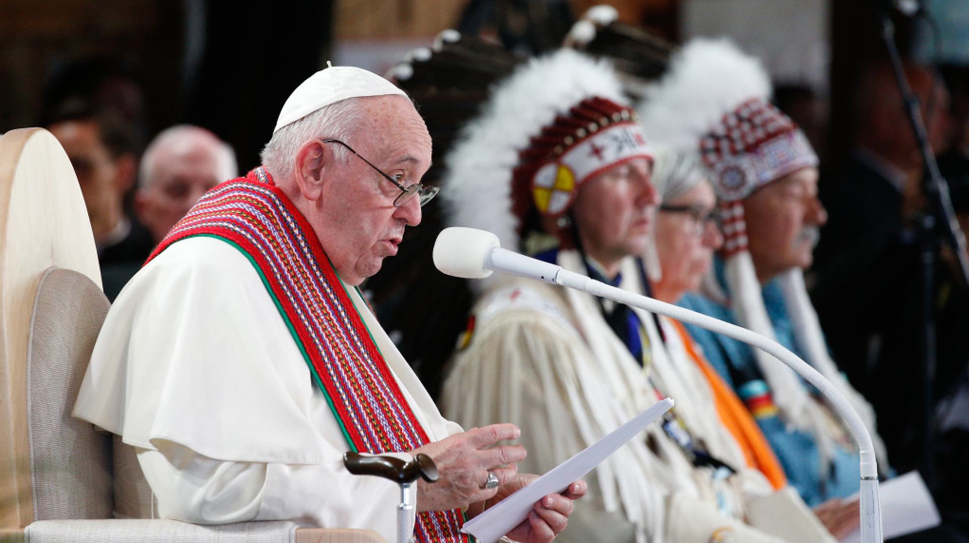 Papst Franziskus sitzt mit Angehörigen der First Nations in Kanada auf einem Podium und spricht in ein Mikrofon