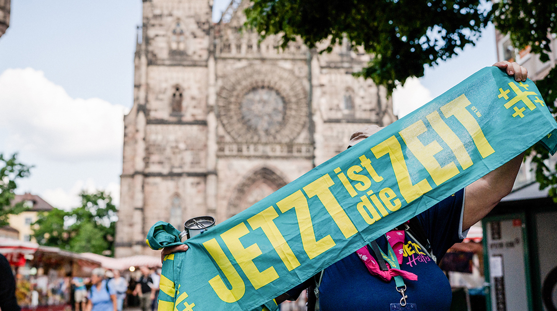 Eine Helferin steht vor der Lorenzkirche und hält einen Schal hoch mit dem Kirchentagsmotto "Jetzt ist die Zeit" auf dem Deutschen Evangelischen Kirchentag am 7. Juni 2023 in Nürnberg.