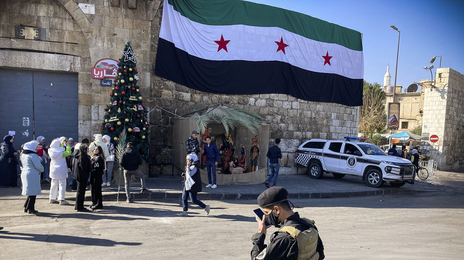 Menschen und Sicherheitskräfte am Bab Touma-Tor, mit einem Weihnachtsbaum und der neuen syrischen Fahne mit drei Sternen, in der Altstadt von Damaskus (Syrien) am 25. Dezember 2024.