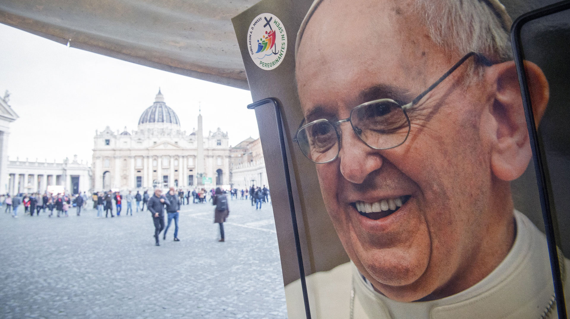 Eine Postkarte mit einem Foto von Papst Franziskus und dem Logo des Heiligen Jahres 2025 in einem Postkartenständer, dahinter Menschen auf dem Petersplatz, am 20. Februar 2025 im Vatikan.