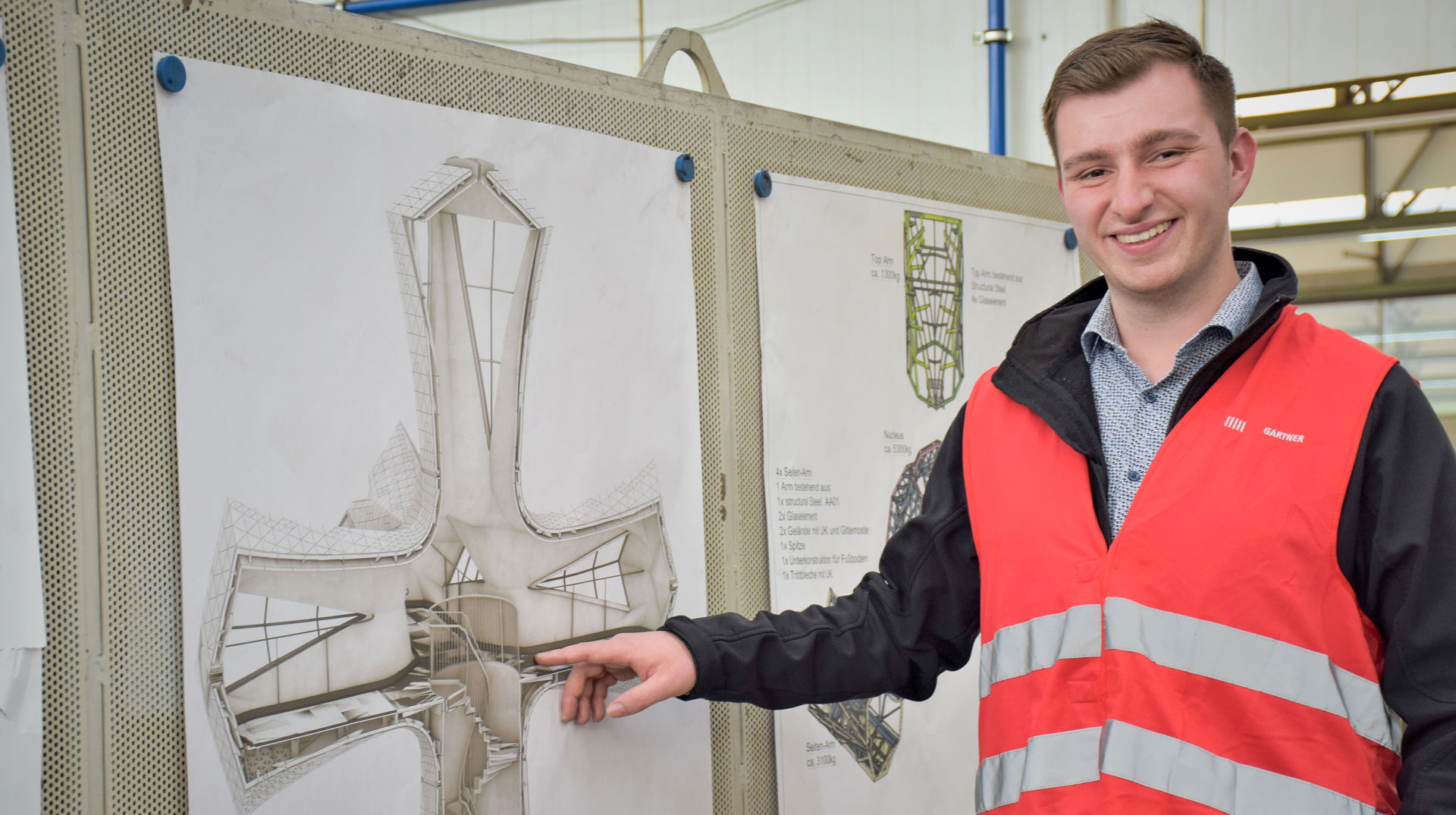 Ein junger Mann zeigt auf den Bauplan eines riesigen, begehbaren Metallkreuzes