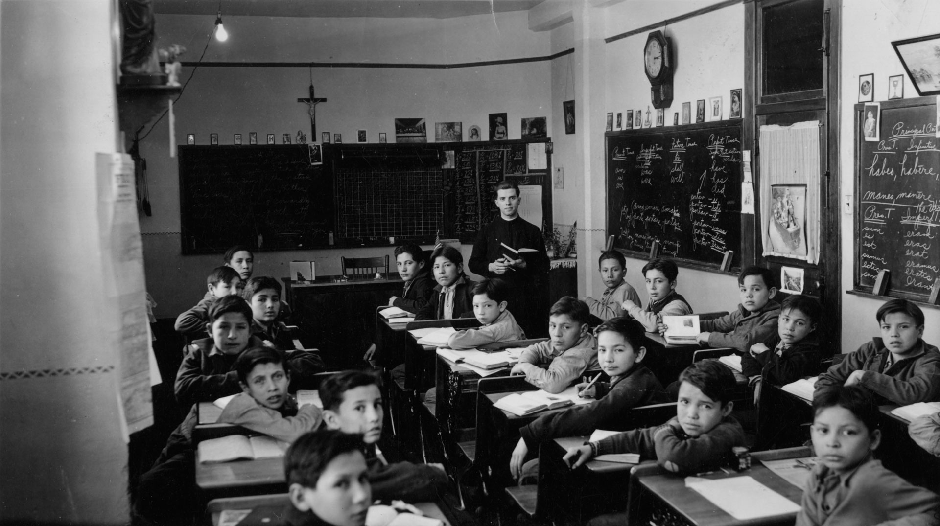 Middle-level classoom with boys and Jesuit teacher, Mr. Alois H. Sommer, S.J., Inscriptions on black boards in image: Pertains to arithmetic and English verb conjugation