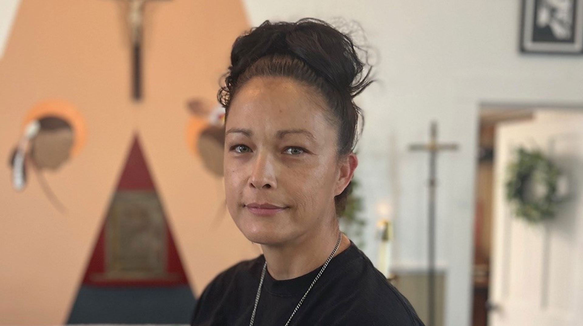 Joyce Tibbitts steht in einem Raum, im Hintergrund sind indigene und christliche Symbole erkennbar