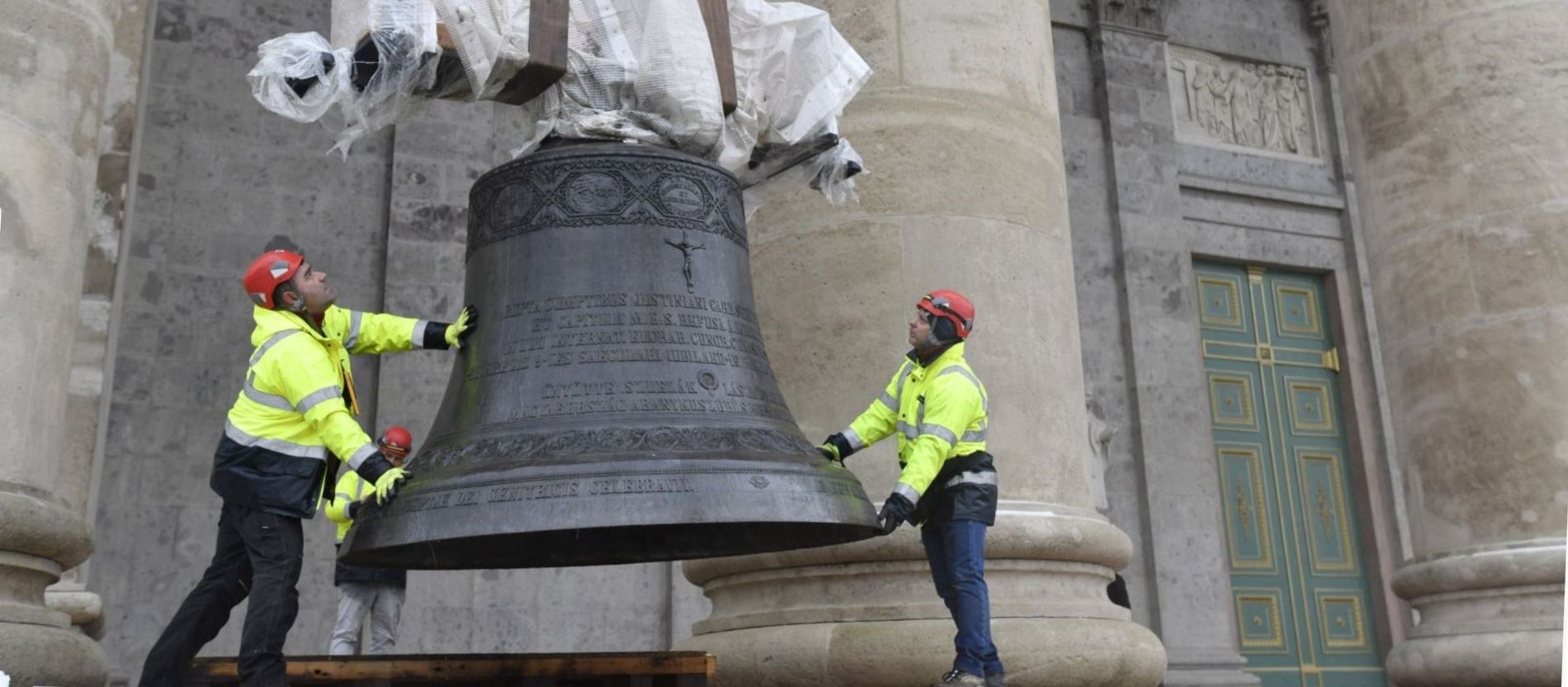 Zwei Männer halten eine Glocke, die an einem Kran hängt. 