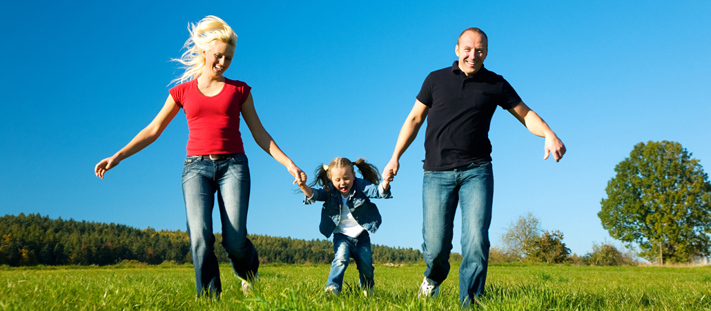 Eine Familie bestehend aus Mutter, Tochter und Vater (von links nach rechts) läuft händchenhaltend eine Wiese entlang.